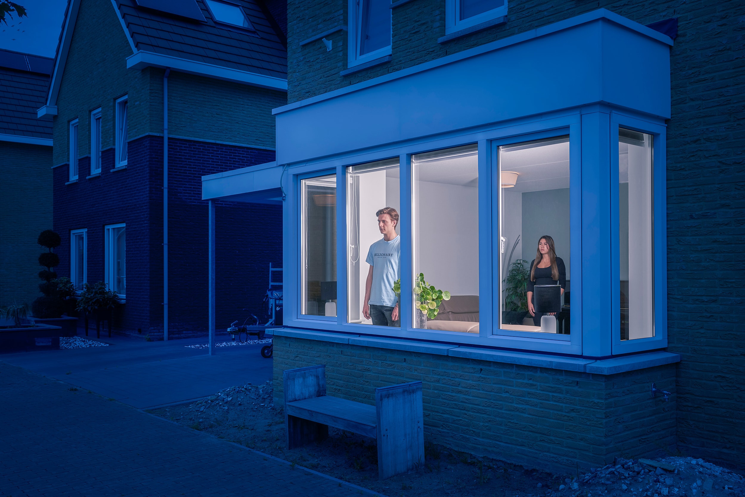 Timo and a woman separated inside a brightly lit home at blue hour